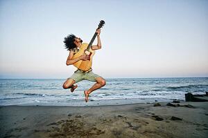 Umělecká fotografie Mixed Race man playing guitar and jumping at beach, Peathegee Inc, (40 x 26.7 cm)